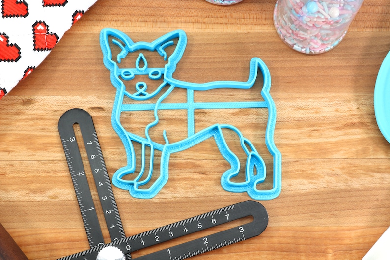 Puede incluir: Cortador de galletas de pl&aacute;stico azul con forma de perro peque&ntilde;o de pie sobre sus cuatro patas. El cortador de galletas est&aacute; sobre una superficie de madera.