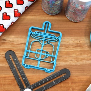 May include: Blue plastic cookie cutter shaped like a coffee maker. The cutter has a handle on top and a cup of coffee inside.