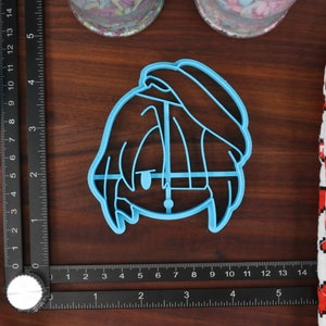 May include: A light blue plastic cookie cutter shaped like a person with short hair and a hat. The cutter is on a wooden surface.