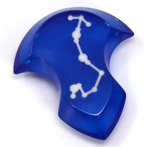 Op de afbeelding: Een blauwe glazen hanger met een wit sterrenbeeld ontwerp. De hanger is gevormd als een halve maan.