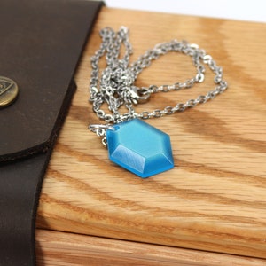 May include: A blue hexagonal pendant necklace with a silver chain. The necklace is resting on a wooden surface.
