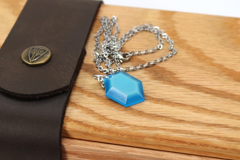 May include: A blue hexagonal pendant necklace with a silver chain. The necklace is resting on a wooden surface.