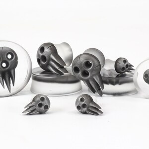 May include: A collection of gray skull-shaped ear gauges in various sizes. The skulls have three eye holes and a jagged mouth. Some are set in clear, circular plugs, while others stand alone. The background is white.
