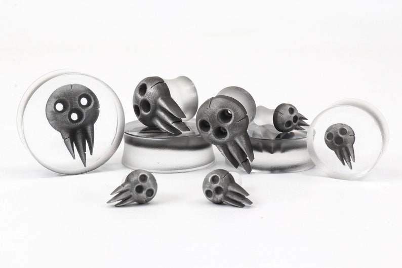 May include: A collection of gray skull-shaped ear gauges in various sizes. The skulls have three eye holes and a jagged mouth. Some are set in clear, circular plugs, while others stand alone. The background is white.