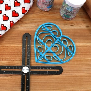 May include: A blue plastic heart-shaped cookie cutter with a decorative design. The cutter is on a wooden surface with a ruler and a rolling pin.