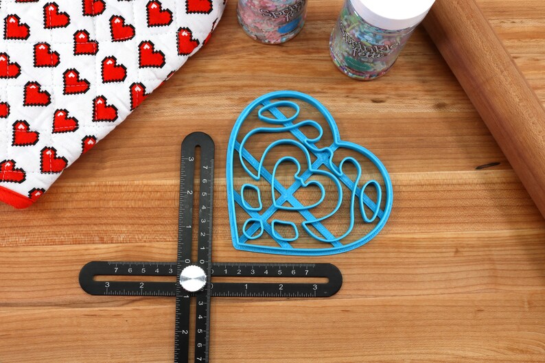 May include: A blue plastic heart-shaped cookie cutter with a decorative design. The cutter is on a wooden surface with a ruler and a rolling pin.