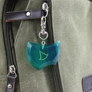 May include: A green and blue resin charm with a stylized letter D inside, attached to a silver key chain. The charm is hanging from a green canvas backpack.