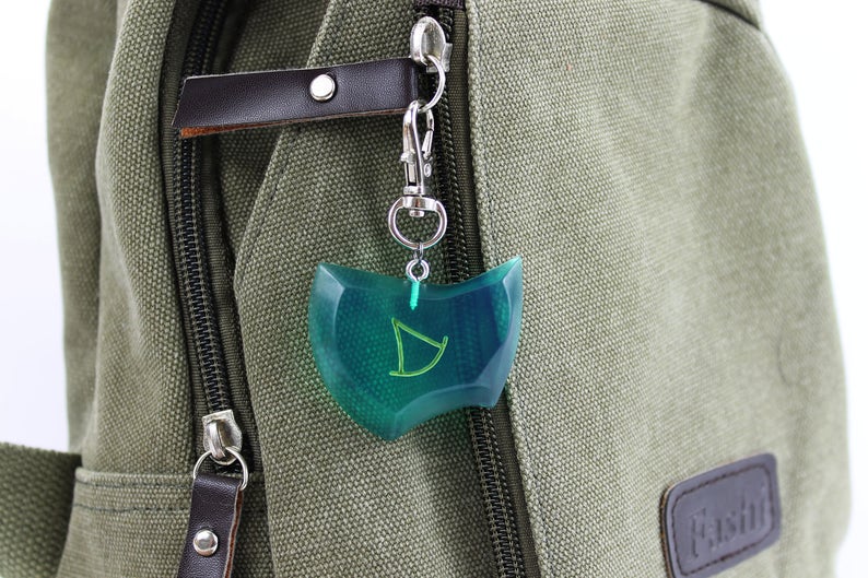 May include: A green and blue resin charm with a stylized letter D inside, attached to a silver key chain. The charm is hanging from a green canvas backpack.