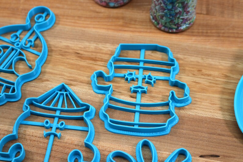 May include: A set of four blue plastic cookie cutters shaped like a snowman, a carousel, a lighthouse, and a Christmas tree. The cutters are on a wooden surface.