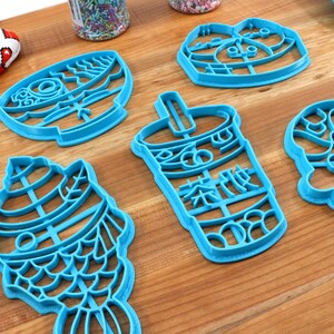 May include: A collection of bright blue cookie cutters in various shapes, including a fish, a bubble tea cup with Chinese characters, and other whimsical designs. The cutters are arranged on a wooden surface.