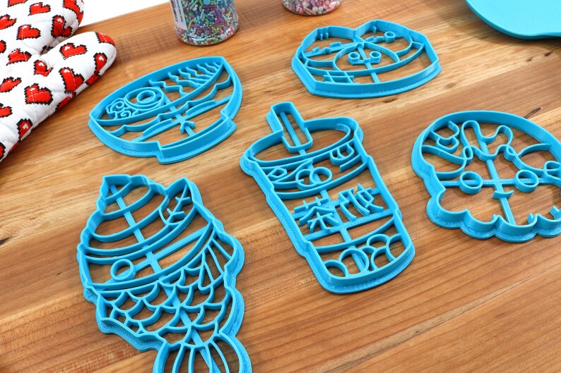 May include: A collection of bright blue cookie cutters in various shapes, including a fish, a bubble tea cup with Chinese characters, and other whimsical designs. The cutters are arranged on a wooden surface.