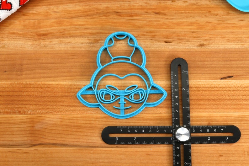 May include: A blue plastic cookie cutter shaped like a cartoon character with a large head, large eyes, and a small mouth. The character has a large, round head with a single, round, raised bump on top. The cookie cutter is approximately 2 inches in diameter.