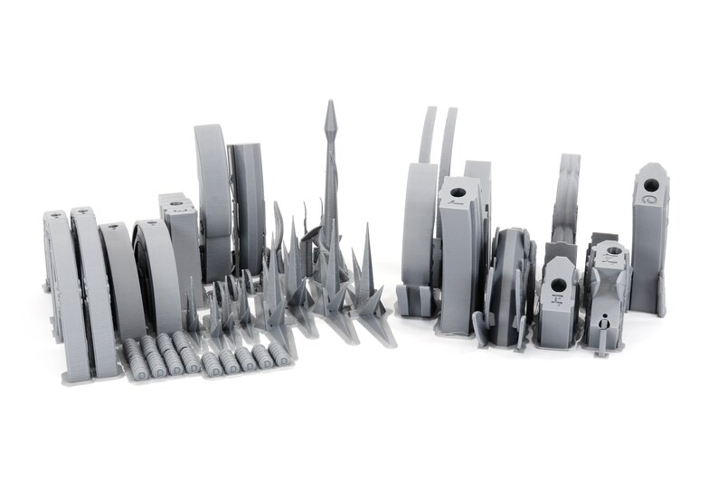 May include: An assortment of gray 3D-printed architectural models, including towers, arches, and cylindrical structures. The collection features various shapes and sizes, showcasing a range of design possibilities. The models are arranged on a white background.