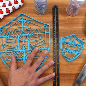 May include: Two blue plastic cookie cutters shaped like the Triforce symbol from the Legend of Zelda video game series. The larger cutter is about 7 inches in diameter and the smaller cutter is about 4 inches in diameter.