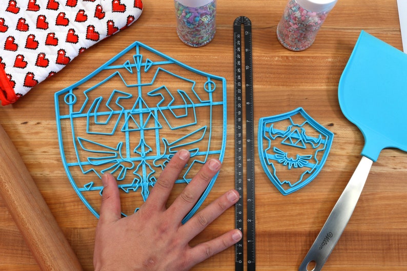 May include: Two blue plastic cookie cutters shaped like the Triforce symbol from the Legend of Zelda video game series. The larger cutter is about 7 inches in diameter and the smaller cutter is about 4 inches in diameter.