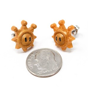 May include: A pair of gold-coloured earrings shaped like a sun with a black face. The earrings are about the size of a ten pence piece.