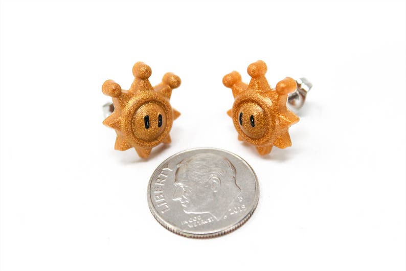 May include: A pair of gold-coloured earrings shaped like a sun with a black face. The earrings are about the size of a ten pence piece.