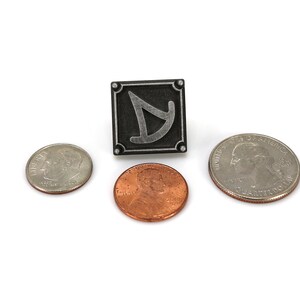 May include: A silver metal pin with a black background and a stylized silver symbol. The pin is surrounded by three US coins, a penny, a dime, and a quarter.