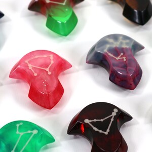 May include: A set of nine colourful resin arrowheads with constellation designs. Each arrowhead is a different colour, including red, green, blue, and purple. The arrowheads are all in focus and are arranged in a semi-circle on a white background.