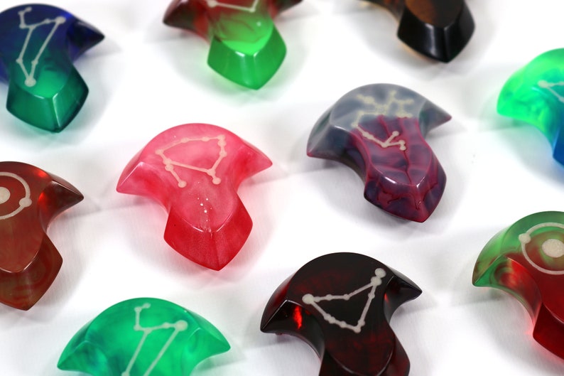 May include: A set of nine colourful resin arrowheads with constellation designs. Each arrowhead is a different colour, including red, green, blue, and purple. The arrowheads are all in focus and are arranged in a semi-circle on a white background.