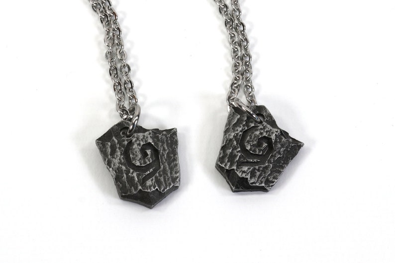 May include: Two silver necklaces with matching pendants. Each pendant is a silver, textured, shield-shaped piece with a stylized, swirling design.