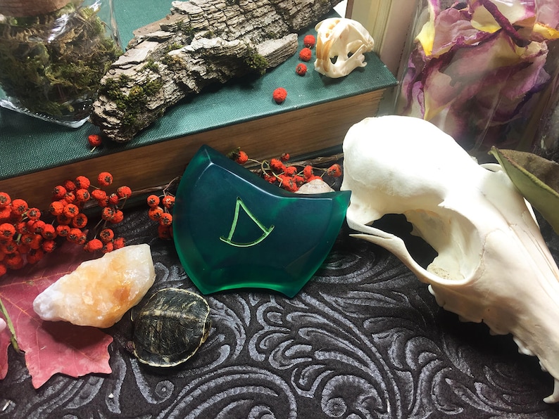 May include: A green resin pendant with a stylized letter D in the center. The pendant is on a dark patterned surface with a small turtle and a piece of orange rock.