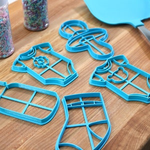 May include: A set of six blue plastic cookie cutters shaped like a baby bottle, a onesie, a sock, a pacifier, and two different onesies. The cutters are on a wooden surface.