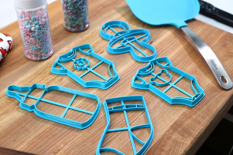 May include: A set of six blue plastic cookie cutters shaped like a baby bottle, a onesie, a sock, a pacifier, and two different onesies. The cutters are on a wooden surface.