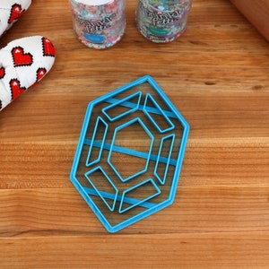 Op de afbeelding: Een blauwe plastic koekjesvormer in de vorm van een geometrisch ontwerp met meerdere lagen. De vormer ligt op een houten oppervlak.