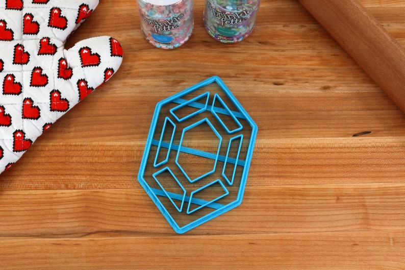 Op de afbeelding: Een blauwe plastic koekjesvormer in de vorm van een geometrisch ontwerp met meerdere lagen. De vormer ligt op een houten oppervlak.
