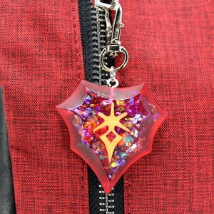 May include: A red and gold shield-shaped charm with a sparkly, iridescent finish. The charm is attached to a silver key chain.