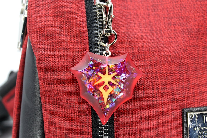 May include: A red and gold shield-shaped charm with a sparkly, iridescent finish. The charm is attached to a silver key chain.
