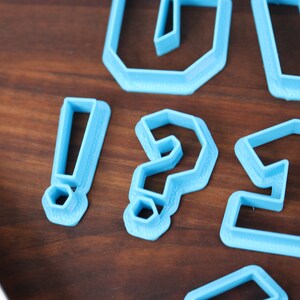 May include: Blue plastic cookie cutters shaped like an exclamation point, question mark, and the number one. The cutters are on a wooden surface.