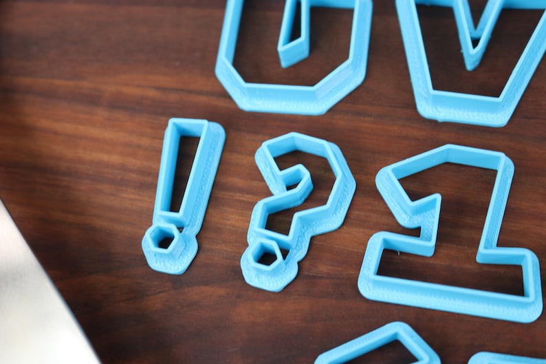 May include: Blue plastic cookie cutters shaped like an exclamation point, question mark, and the number one. The cutters are on a wooden surface.