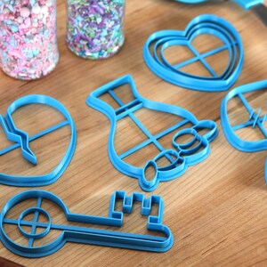 May include: A set of blue plastic cookie cutters in the shapes of a key, a heart, a hand holding a heart, a broken heart, and a dress. The cutters are on a wooden surface.