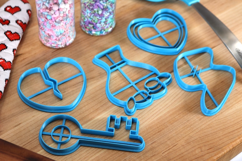 May include: A set of blue plastic cookie cutters in the shapes of a key, a heart, a hand holding a heart, a broken heart, and a dress. The cutters are on a wooden surface.