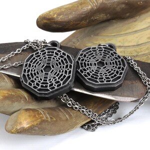 May include: Two silver-toned necklaces with geometric pendant charms. The charms are octagonal and feature a detailed, intricate pattern. The necklaces are displayed on a wooden surface.
