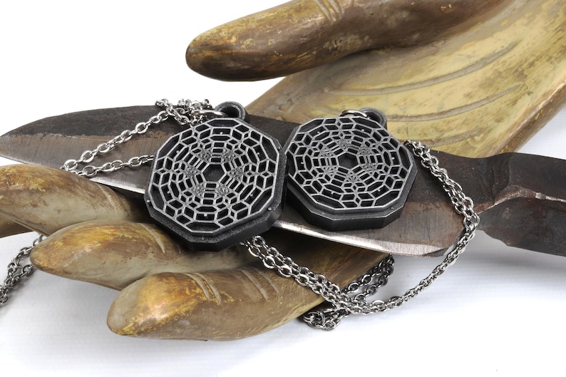 May include: Two silver-toned necklaces with geometric pendant charms. The charms are octagonal and feature a detailed, intricate pattern. The necklaces are displayed on a wooden surface.