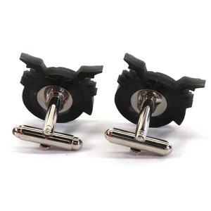 May include: A pair of black cufflinks with a silver metal backing. The cufflinks feature a stylized design of a record player.