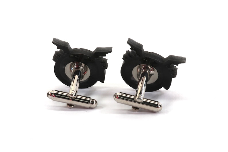 May include: A pair of black cufflinks with a silver metal backing. The cufflinks feature a stylized design of a record player.