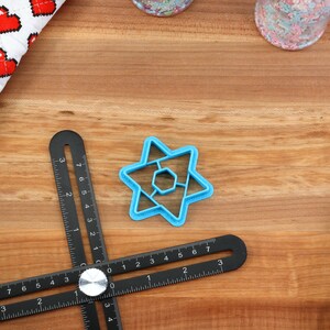 May include: A blue plastic cookie cutter shaped like a six-pointed star with a smaller six-pointed star inside. The cookie cutter is on a wooden surface with a black ruler and a white and red fabric with hearts.