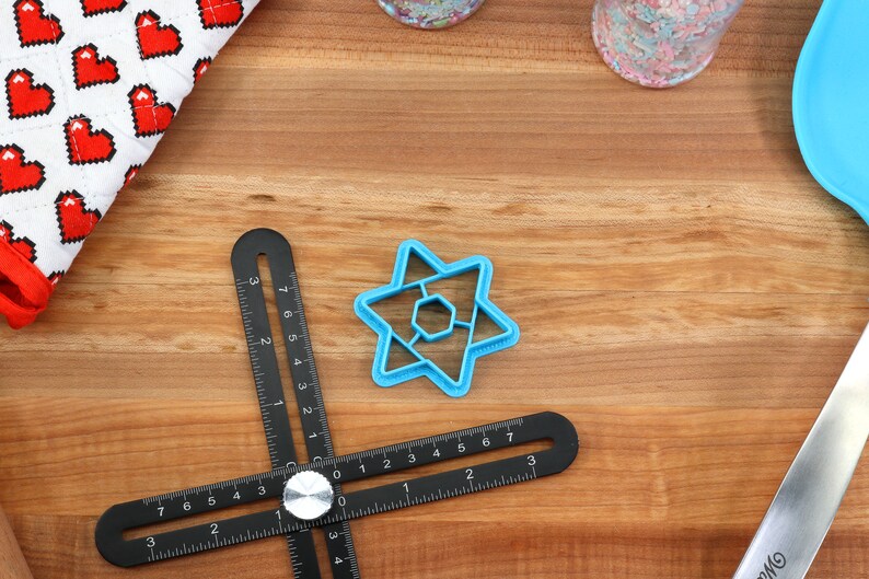 May include: A blue plastic cookie cutter shaped like a six-pointed star with a smaller six-pointed star inside. The cookie cutter is on a wooden surface with a black ruler and a white and red fabric with hearts.