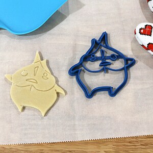 May include: Two cookie cutters, one blue and one beige, shaped like a cartoon character with a surprised expression. The blue cutter is on a white surface, and the beige cutter is on a white surface with a blue spatula.