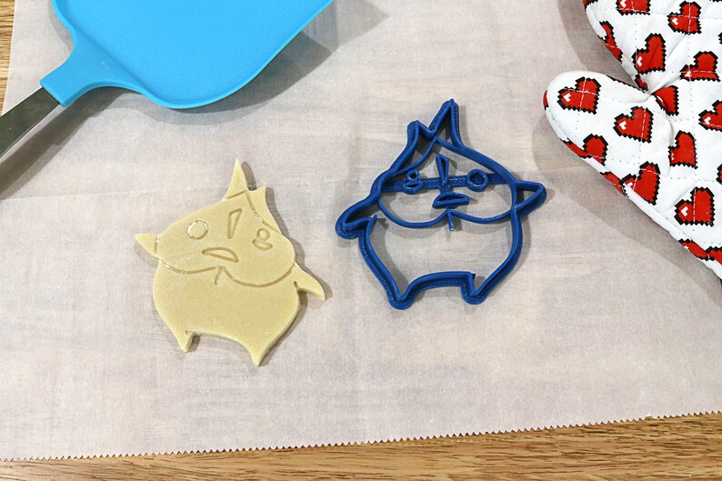 May include: Two cookie cutters, one blue and one beige, shaped like a cartoon character with a surprised expression. The blue cutter is on a white surface, and the beige cutter is on a white surface with a blue spatula.