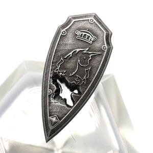 May include: A silver shield with a unicorn design and a crown at the top. The shield is slightly damaged, with a small hole in the center.