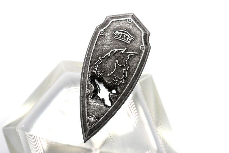 May include: A silver shield with a unicorn design and a crown at the top. The shield is slightly damaged, with a small hole in the center.