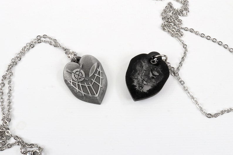 May include: Two silver heart-shaped pendants on chains. One pendant has a detailed design of a target with lines radiating out. The other pendant is black and has a textured surface.