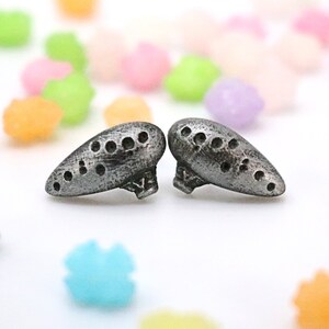 Pu&ograve; includere: Due ocarine argentate e stilizzate con pi&ugrave; fori sono esposte su una superficie bianca. Le ocarine sono in primo piano, con caramelle colorate a forma di stella sparse sullo sfondo. Le ocarine hanno un aspetto metallico strutturato.