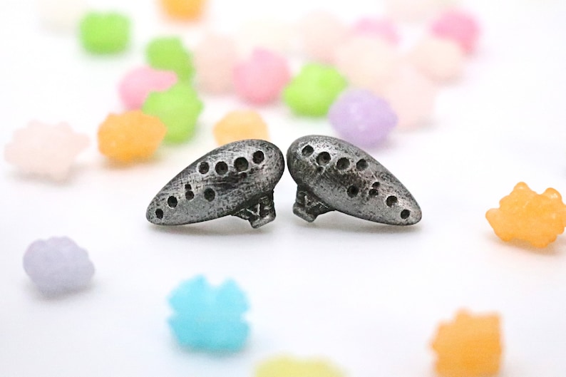 Pu&ograve; includere: Due ocarine argentate e stilizzate con pi&ugrave; fori sono esposte su una superficie bianca. Le ocarine sono in primo piano, con caramelle colorate a forma di stella sparse sullo sfondo. Le ocarine hanno un aspetto metallico strutturato.