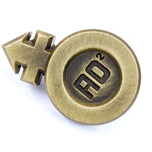 May include: A gold-colored metal pin with a stylized arrow and a circle containing the text "AD&sup2;".